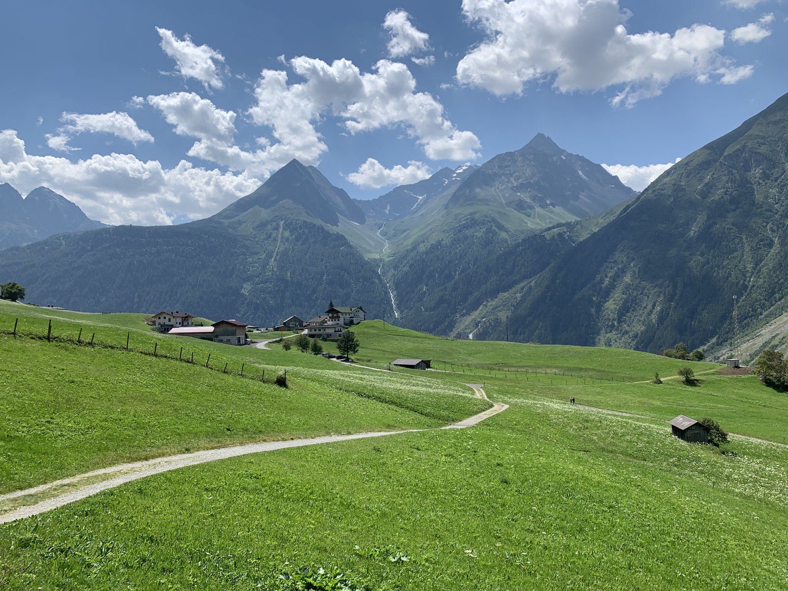 Een wandelpad door Alpenweide leidt naar een houten schuur en bergpiek op de achtergrond